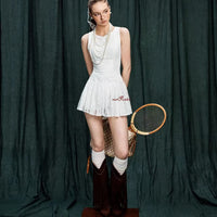 Woman in a white tennis outfit with a vintage racket against a dark green curtain.