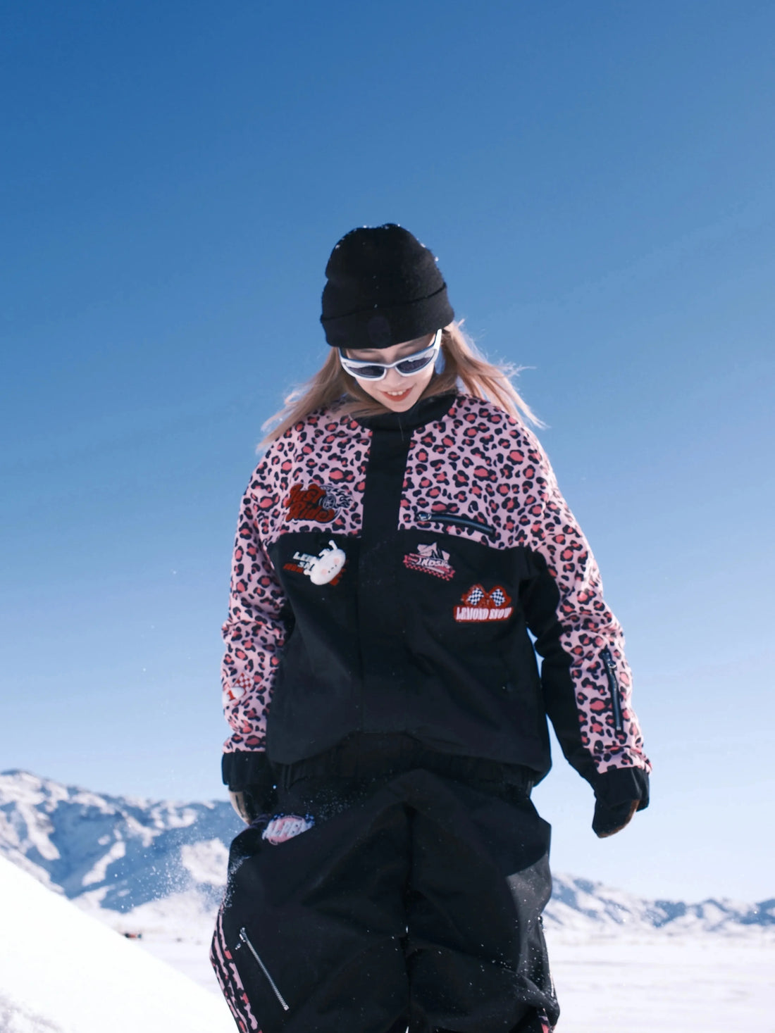 Person in leopard print jacket and black pants standing on a snowy slope with a clear blue sky.