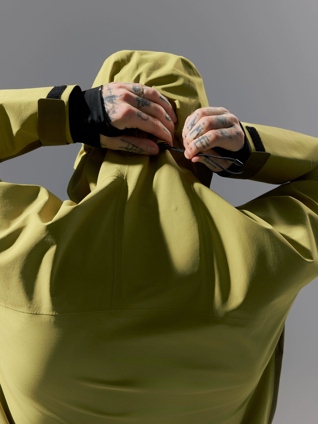 Person wearing a light green jacket with tattoos on arms against a gray background
