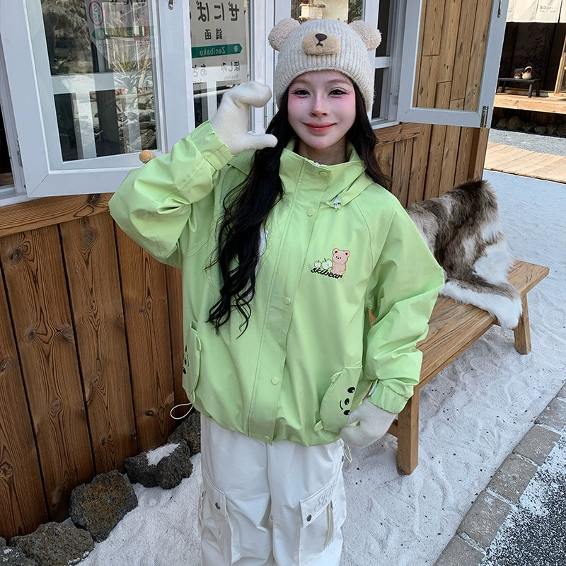 Person wearing a light green snowboarding jacket with a bear design, standing outdoors in front of a wooden building.