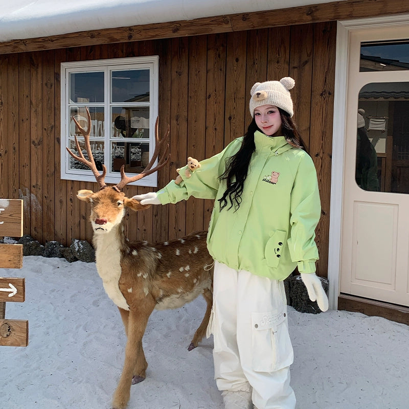 Person in green snowboarding jacket and white pants interacting with a deer in front of a wooden building.