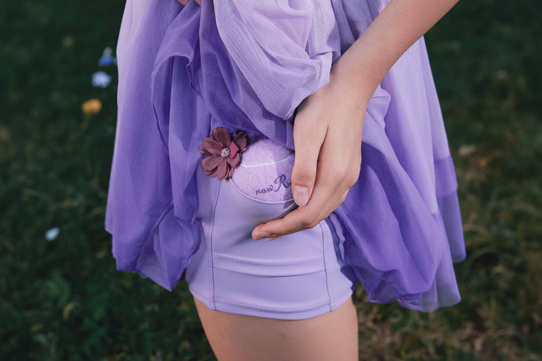 Person wearing a purple dress with a floral detail, holding a matching bag against a blurred green background.