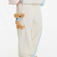 Person wearing a light blue jacket and cream pants holding a small teddy bear with a blue scarf.