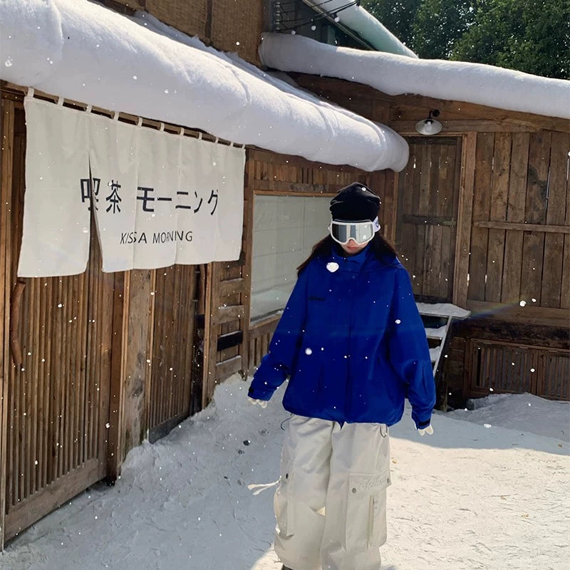 Person in blue snowboarding jacket and white pants standing in front of a wooden building with snow on the roof.