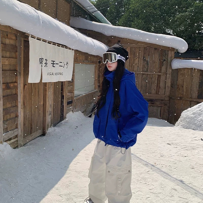 Person in blue jacket and white pants standing in front of a wooden building with snow on the roof.