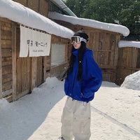 Person in blue jacket and white pants standing in front of a wooden building with snow on the roof.