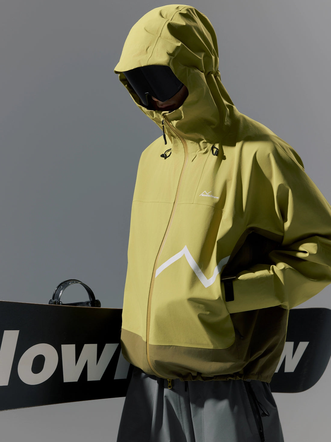 Person wearing a green jacket with a brand logo, holding a black snowboard with white text.