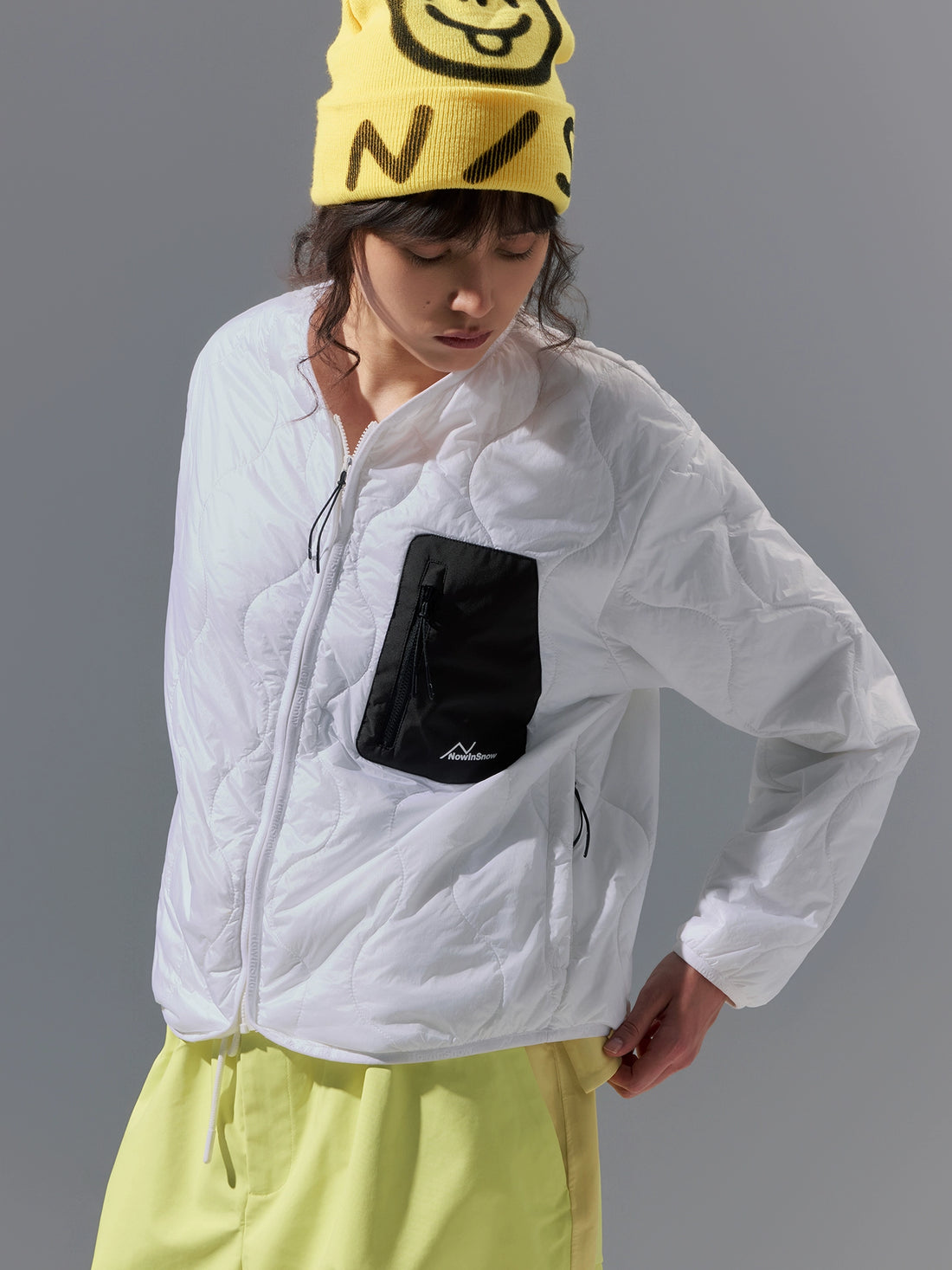 Person wearing a white jacket with a black pocket and yellow pants, standing against a gray background.