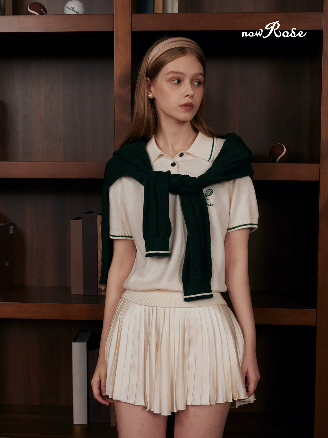 Woman wearing a white blouse with a green scarf and pleated skirt in front of a wooden shelf.
