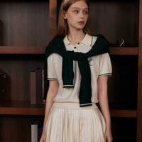 Woman wearing a white blouse with a green scarf and pleated skirt in front of a wooden shelf.
