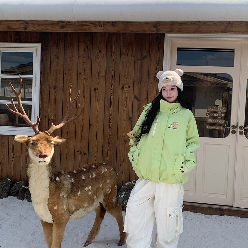 Person in a green jacket and white pants standing next to a deer in front of a wooden cabin.