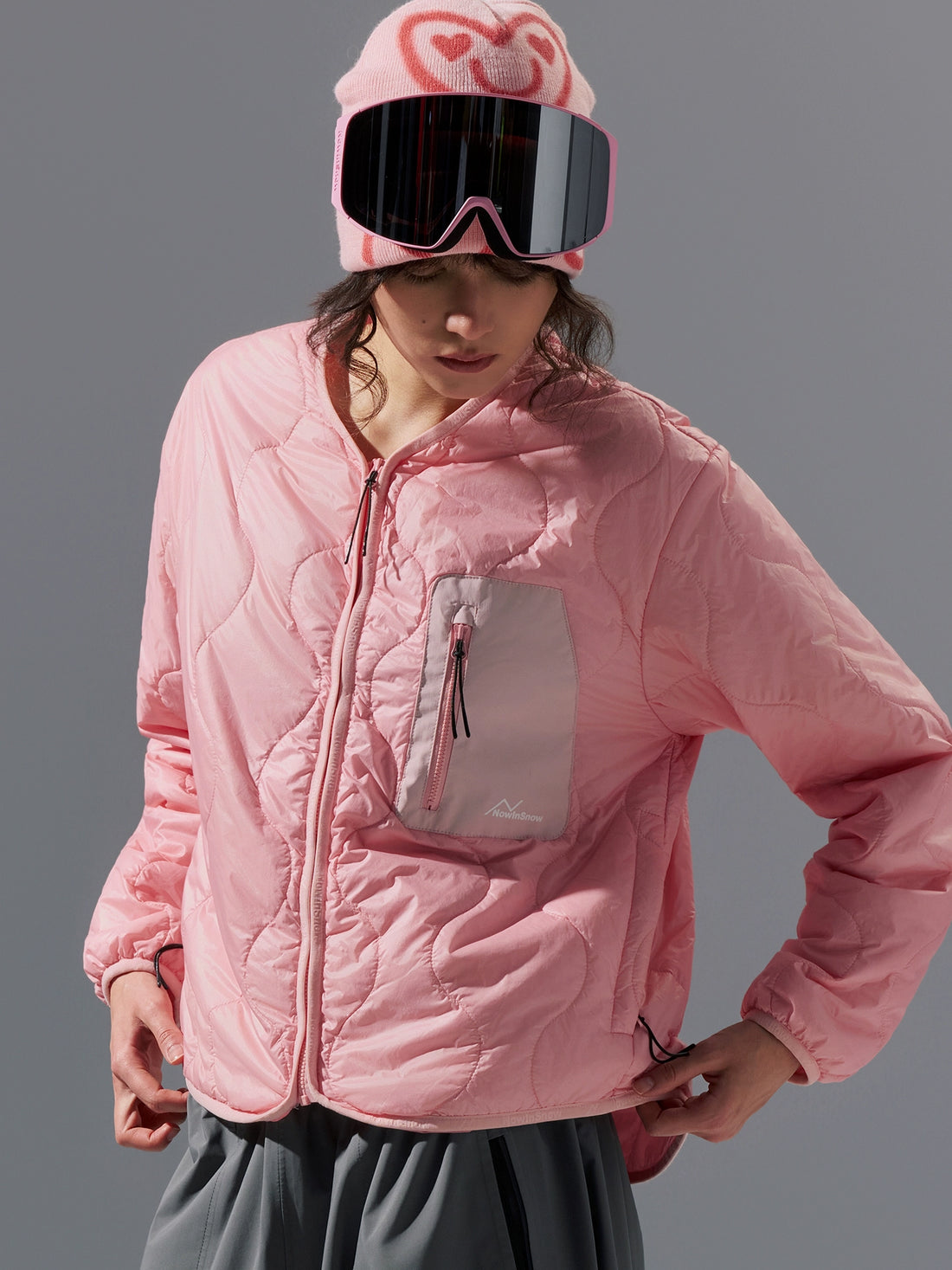 Person wearing a pink quilted jacket, gray pants, and a matching pink hat with goggles on a gray background