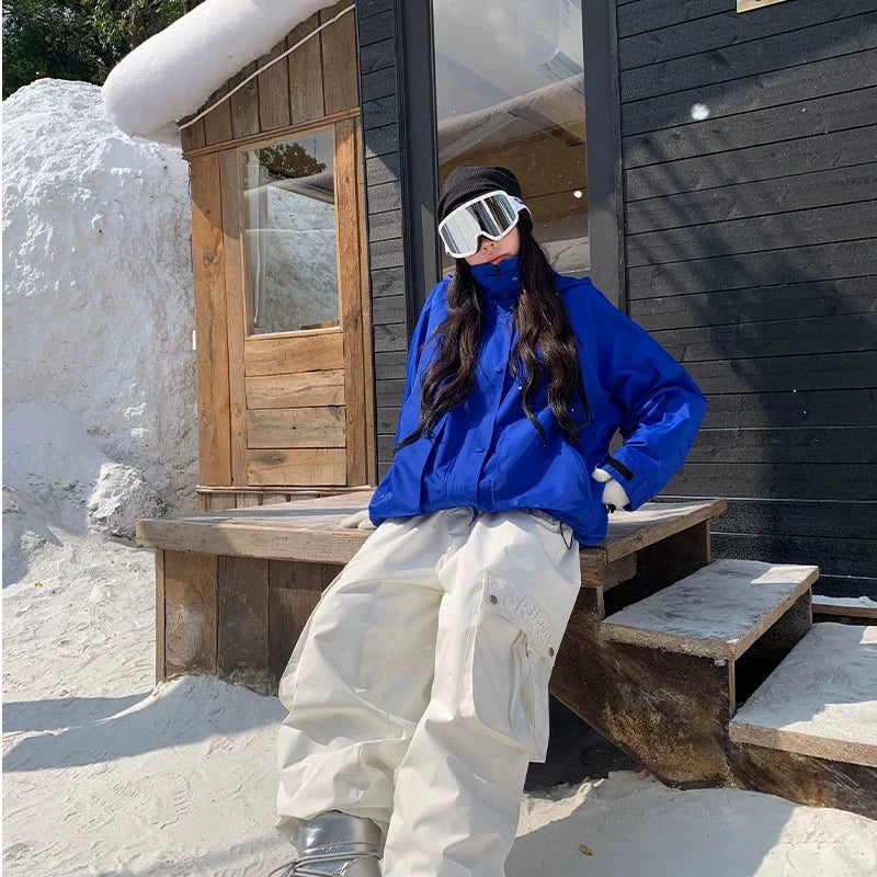 Person in blue snowboarding jacket and white pants sitting on a wooden bench outside a snow-covered cabin.