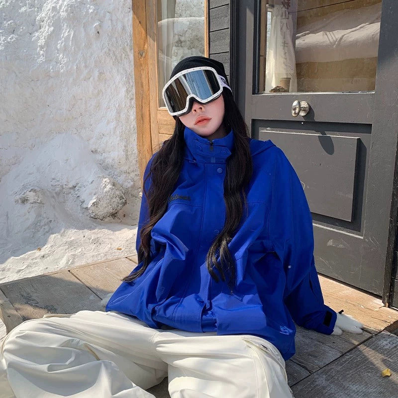 Person wearing a blue snowboarding jacket and white pants sitting on a stone surface with a wooden door in the background.
